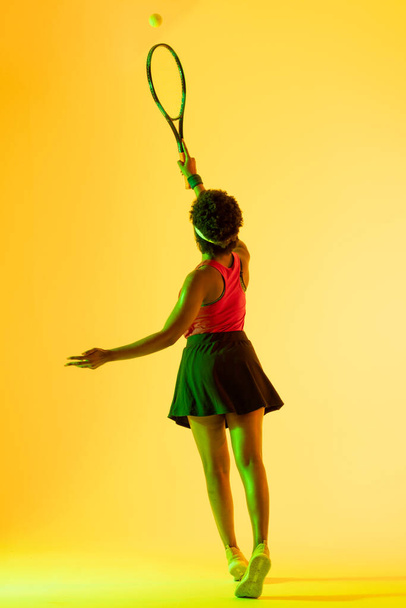Vertical image of back view of african american female tennis player in yellow lighting. Sport, movement, fitness and active lifestyle concept. - Foto, Imagem
