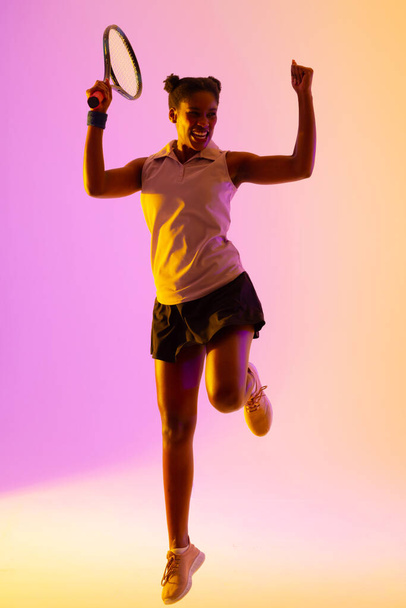 Vertical image of successful african american female tennis player in violet and yellow lighting. Sport, movement, fitness and active lifestyle concept. - Foto, Imagen