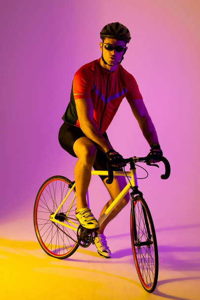 Portrait of caucasian male cyclist with bicycle over pink lighting. Sport, movement, training and active lifestyle concept. - Photo, Image