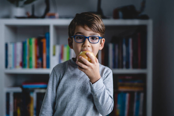 Портрет одного маленького белого мальчика в очках, смотрящего в камеру и поедающего яблоко дома - Фото, изображение