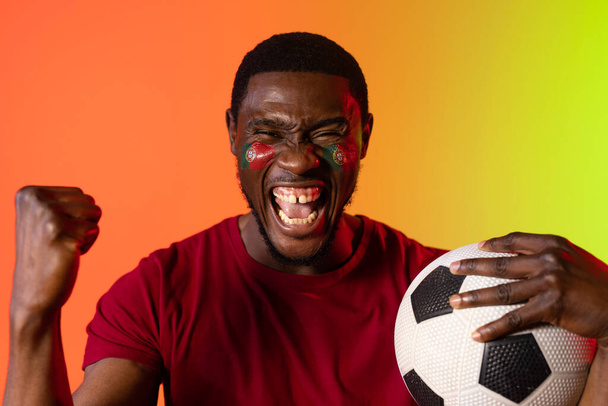 Portrait of african american male supporter with flag of portugal on cheeks over orange lighting. Sport, movement, training and active lifestyle concept. - Fotografie, Obrázek
