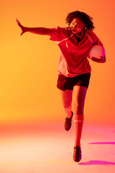 African american female rugby player with arm wide and rugby ball over neon pink lighting. Sport, movement, training and active lifestyle concept. - Valokuva, kuva
