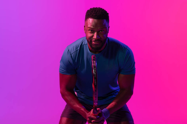 Image of happy african american male tennis player in violet and pink neon lighting. Sport, movement, fitness and active lifestyle concept. - Φωτογραφία, εικόνα