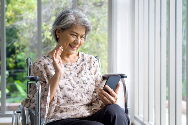 Asian gray hair old woman waving her hand via remote communication through mobile phone while sitting on wheelchair in front of corridor with glass wall. - 写真・画像