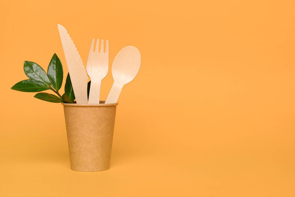 Natural wooden bamboo cutlery set in paper cup - biodegradable and compostable cutlery and tableware concept. Bamboo cutlery against orange background with copy space. Environmental conservation - 写真・画像