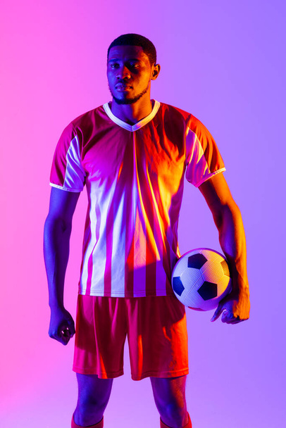 African american male soccer player with football over neon pink lighting. Sport, movement, training and active lifestyle concept. - Foto, afbeelding