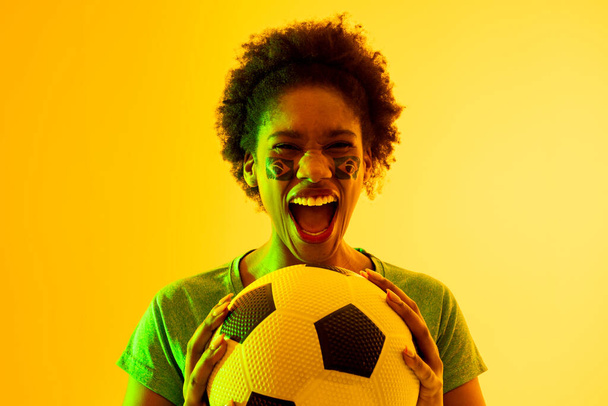 Image of african american female soccer fan with flag of brazil cheering in yellow lighting. Sport, fans, cheering and emotions concept. - Foto, Bild