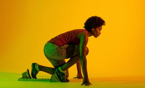 Image of african american female athlete preparing for run in yellow lighting. Sport, movement, fitness and active lifestyle concept. - Foto, Imagen