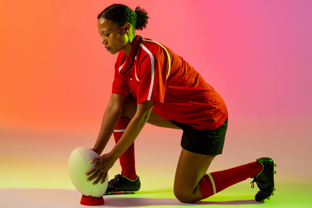 African american female rugby player crouching with rugby ball over neon pink lighting. Sport, movement, training and active lifestyle concept. - Photo, image