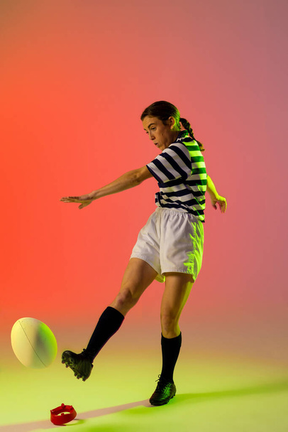 Caucasian female rugby player kicking rugby ball over neon pink lighting. Sport, movement, training and active lifestyle concept. - Zdjęcie, obraz
