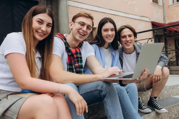 Study together is fun, teamwork, team building concept. Four happy students are sitting near college building and holding books, devices, wearing casual smart, smiling, on a nice summer - Photo, Image