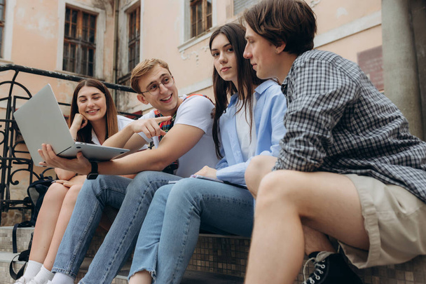 Study together is fun, teamwork, team building concept. Four happy students are sitting near college building and holding books, devices, wearing casual smart, smiling, on a nice summer - Foto, imagen