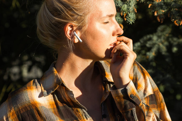 Attraktive kaukasische Blondine im karierten Hemd, die im Stadtpark Musik mit drahtlosen Kopfhörern hört. Junge nachdenkliche Frauen nutzen und genießen Kopfhörer-App. Menschen und Gadgets-Konzept. - Foto, Bild