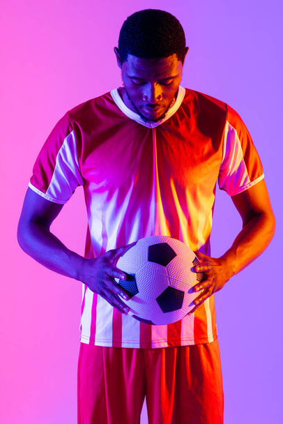 African american male soccer player with football over neon pink lighting. Sport, movement, training and active lifestyle concept. - Фото, изображение