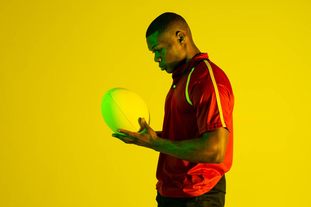 African american male rugby player with rugby ball over yellow lighting. Sport, movement, training and active lifestyle concept. - Foto, Imagem
