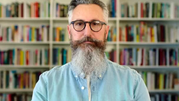 University lecturer and literature professor standing in front of a library bookshelf. Portrait of a serious and intelligent intellectual professor, researcher or author with a grey beard and glasses. - Felvétel, videó