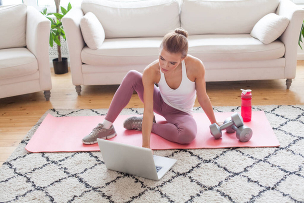 Подходит симпатичная модель в спортивной одежде, сидящая на коврике с помощью ноутбука и готовящаяся к тренировке дома - Фото, изображение