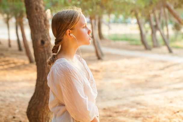 Задоволена щаслива молода дівчина-підліток на відкритому повітрі в красивому сонячному парку, слухаючи музику з навушниками
. - Фото, зображення