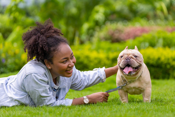 Afro-Amerikaanse vrouw speelt met haar franse bulldog puppy terwijl liggend in het gras gazon na het hebben van ochtend oefening in het park - Foto, afbeelding