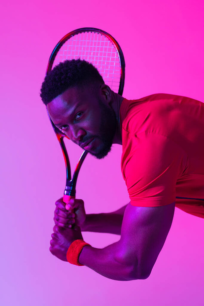 Vertical image of african american male tennis player in neon pink lighting. Sport, movement, fitness and active lifestyle concept. - Fotografie, Obrázek