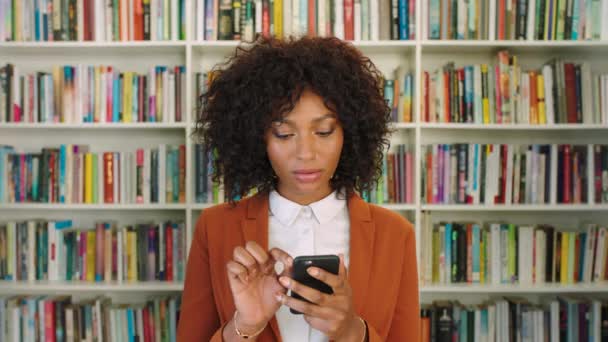 Author and blogger reading reviews online. Young woman browsing and texting on phone with bookshelf in the background. Happy entrepreneur, lecturer or academic researcher scrolling social media apps. - Filmati, video