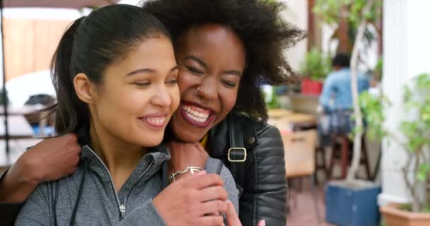 Two best friends bonding and hugging during a social visit. Happy female friends hugging outside in greeting. .Excited african girl embracing her smiling friend from behind in surprise meeting - Πλάνα, βίντεο