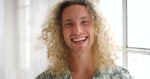 Closeup portrait of a happy man laughing and looking cheerful inside a bright room. Face of a handsome trendy male smiling beside a sunny window at home. A stylish young guy with blonde curly hair. - Footage, Video