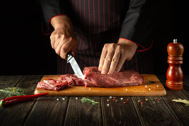 Szef kuchni kroi surowe świeże mięso wołowe na desce do krojenia przed pieczeniem lub grillowaniem. Środowisko pracy w kuchni restauracji lub hotelu. - Zdjęcie, obraz