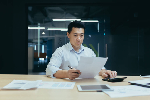 Young Asian male accountant working with documents and calculator. Sitting at a desk with a laptop in a modern office - Foto, Imagem