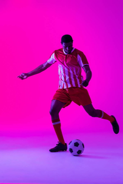 African american male soccer player kicking football over neon pink lighting. Sport, movement, training and active lifestyle concept. - Zdjęcie, obraz