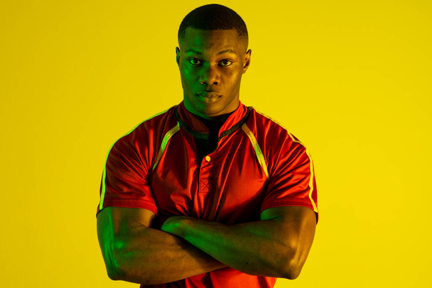 Portrait of african american male rugby player over yellow lighting. Sport, movement, training and active lifestyle concept. - Foto, afbeelding