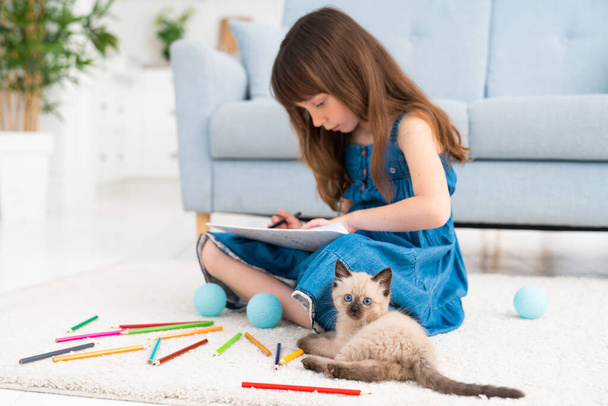Child and kitten. A cute little girl sits on the floor at home and draws with colored pencils. A little fluffy cat is playing nearby. High quality photo - Фото, изображение