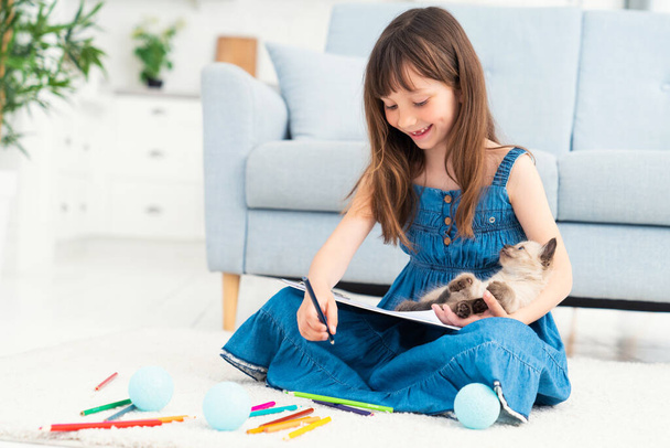 A cute girl sits on the floor at home, draws with colored pencils and plays with a little kitten. The concept of children and love for animals. High quality photo - Фото, изображение