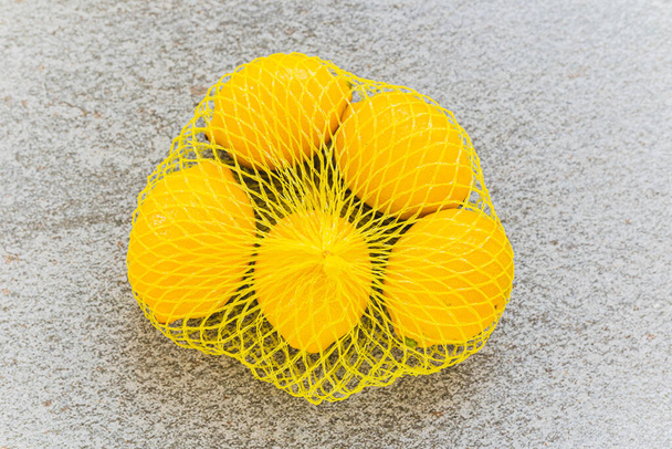 Желтые яркие лимоны в экологической сетке желтого цвета на размытом каменном фоне сверху. Яркий цвет цитрусовых на сером фоне - Фото, изображение