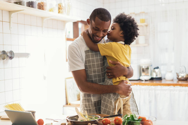 Sweet moments of fatherhood concept. Cute African American boy embracing and kiss on cheek his father while prepare spaghetti meal together in kitchen at home. Happy Black family activity together. - 写真・画像