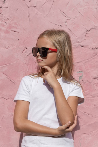 A preteen girl with blond hair poses for the camera against a pink wall. - Foto, Imagen