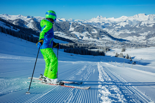 Preteen boy stand on alpine ski look down on fresh skiing track with mountains peak on background on sunny morning - Photo, Image