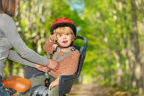 Close-up portrait of a little blond boy point finger sitting in the bicycle seat while mother attach belt and red helmet in the park - Фото, изображение