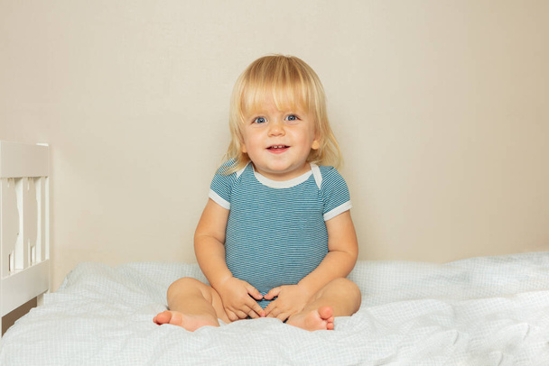 Portrait of a happy smiling blond toddler boy with open mouth sit on the bed in nursery - Zdjęcie, obraz