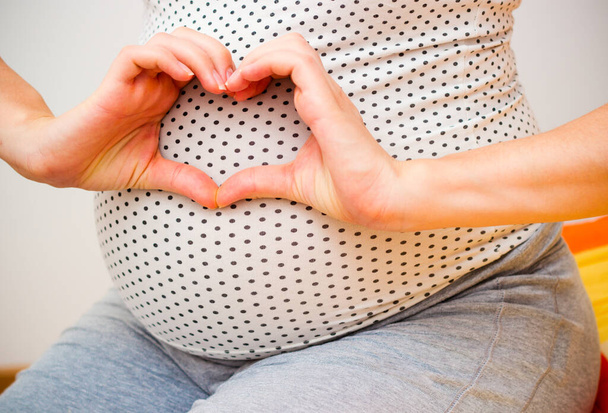 Feliz mujer embarazada haciendo un gesto de corazón. Toma de la mano en el vientre en los interiores de casa. - Foto, imagen
