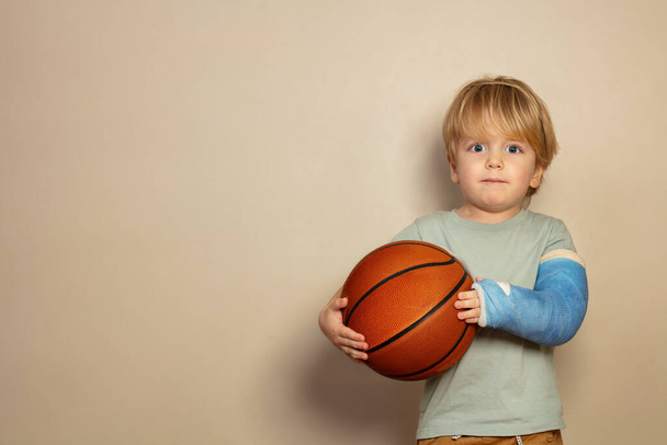Little blond boy hold big orange basketball ball in hands with broken hand in blue plaster cast - Foto, Bild