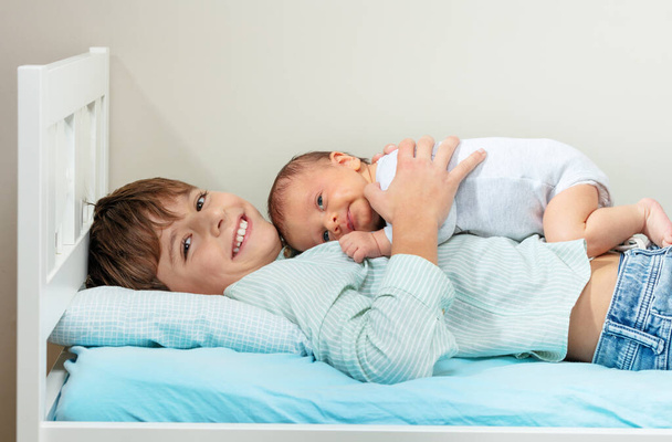 Laughing smiling young positive boy lay in the bed hugging newborn sister - Fotó, kép