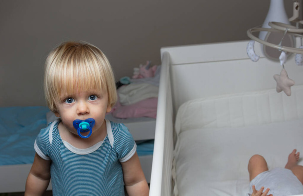 Little blond boy with pacifier stand near the crib of newborn baby - Φωτογραφία, εικόνα