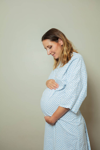 Portrait of a young pregnant woman in hospital robe holding big belly profile view - Photo, image
