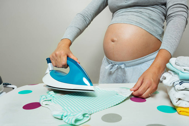 Pregnant woman ironing newborn baby clothes preparing for birth of a child with close-up on Iron - 写真・画像