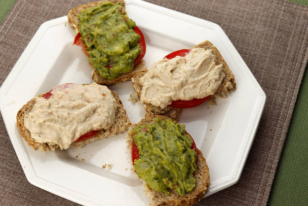 Four half slices of sprouted grain bread, two of them with garlic hummus and a tomato slice and two with tomato slices and guacamole. Bread with guacamole, garlic hummus and tomato slices on a plate. - Photo, Image