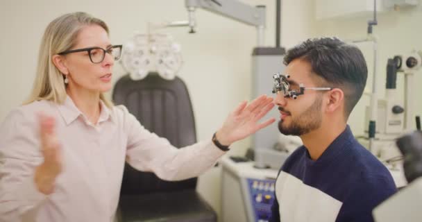 Optometrist doet een oogtest op een patiënt in een kliniek. Vrouwelijke opticien beoordeelt de visie van een jonge man met een phoropter. Oogverzorger helpt zicht te corrigeren met voorgeschreven lenzen. - Video