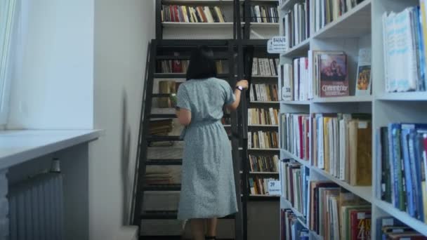 gelukkige en dromerige vrouw leest een boek in de bibliotheek, kiest boeken uit de kast. leren in de bibliotheek voor studenten. - Video