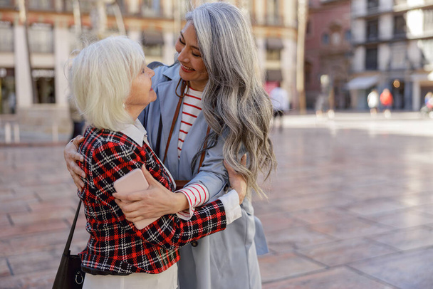 Cheerful aged woman embracing female friend with long gray hair during meeting on modern city street - Photo, Image