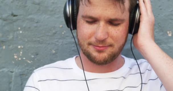 4k video footage of a handsome young man listening to music on his headphones while standing against a brick wall. - Πλάνα, βίντεο